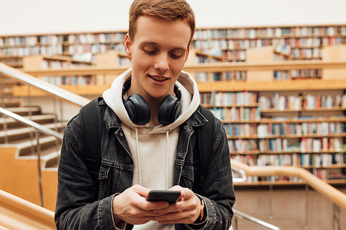 Guy with ginger hair typing on a smartphone in a library. Student with headphones in college.