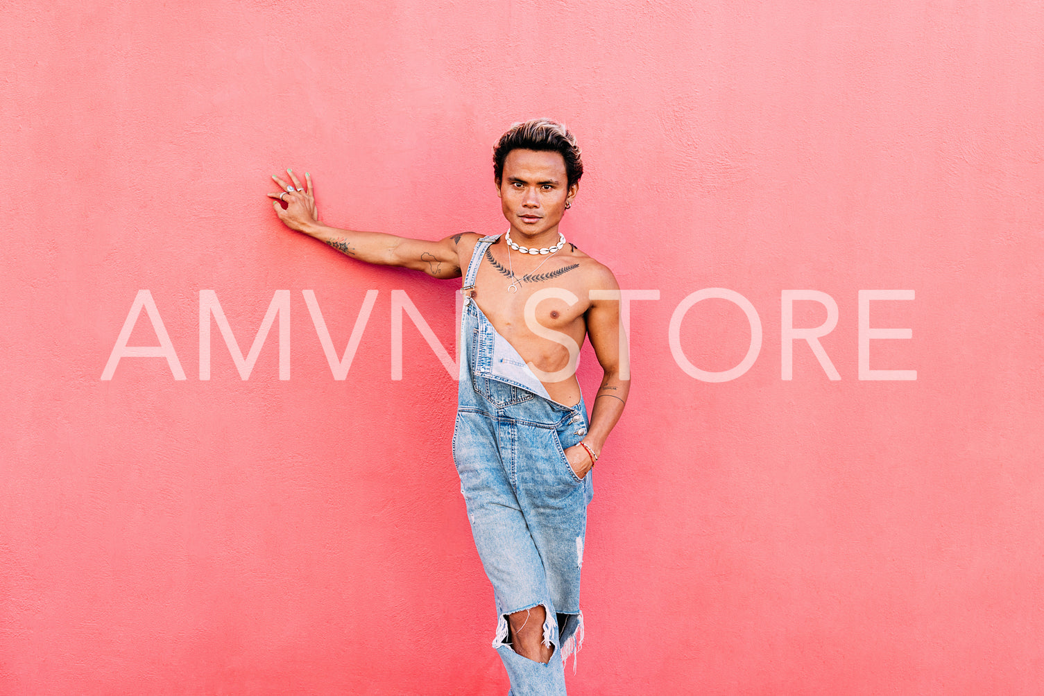 Young handsome man wearing jeans overall leaning pink wall looking at camera