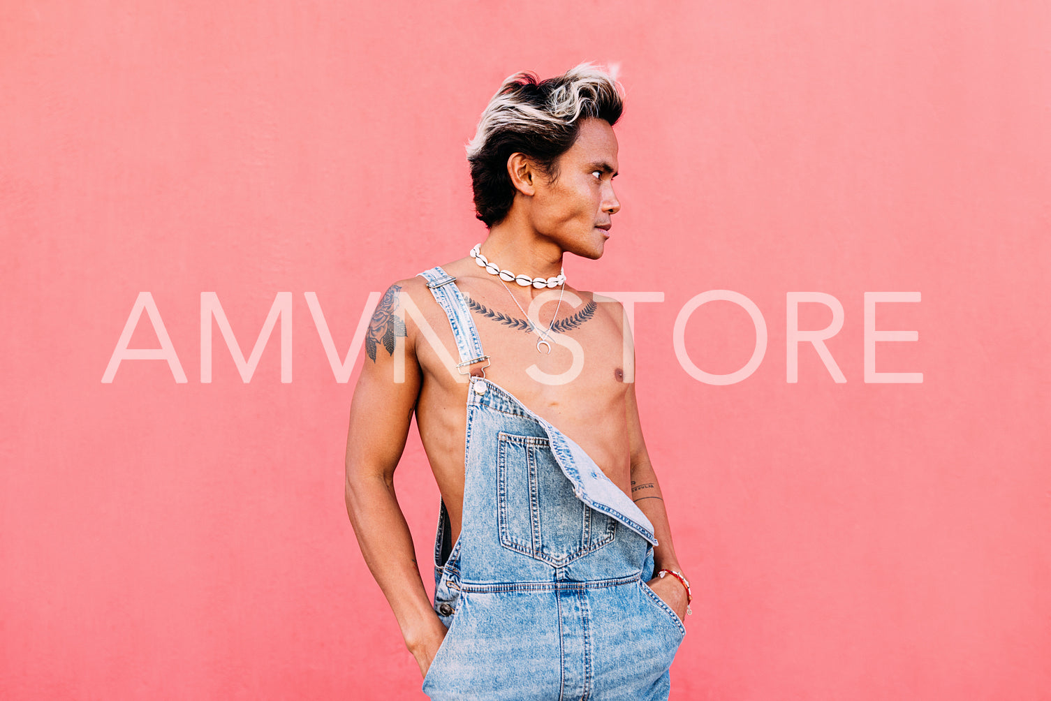 Young stylish guy in denim overalls standing outdoors at pink wall and looking away