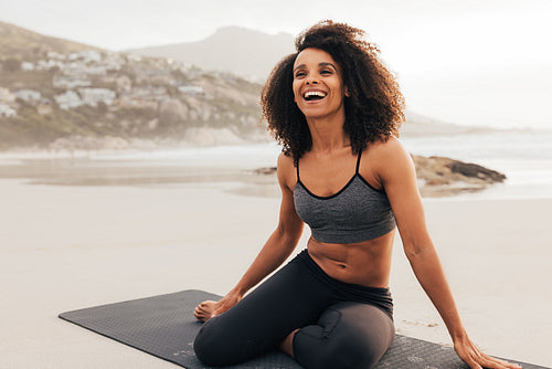 Laughing woman sitting on a beach on mat. Cheerful female relaxing after yoga training.