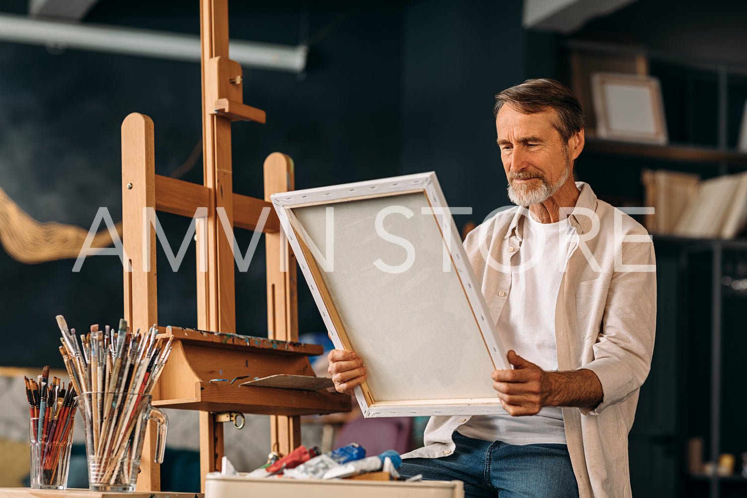 Senior painter sitting in his creative studio and holding a canvas with a picture	