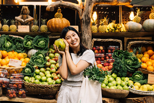 Portrait of a smiling Asian girl holding a fruit and looking at the camera. Young female with a bag at an outdoor market choosing fruits.