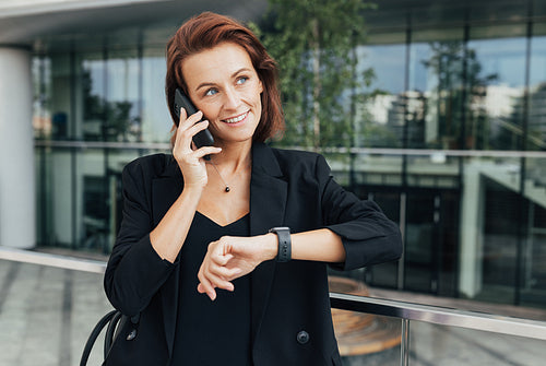 Middle-aged businesswoman talking on mobile phone and looking away. Smiling woman in black formal wear sets a time for a meeting