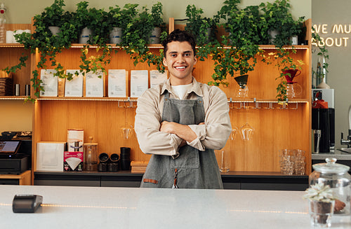 Confident male barista in an apron at the counter. Bartender with crossed hands looking at the camera and smiling.