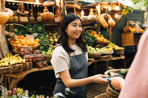 Outdoor market owner receiving payment from the buyer. Asian woman in an apron holding a pos terminal looking at a customer at a local farmer market.