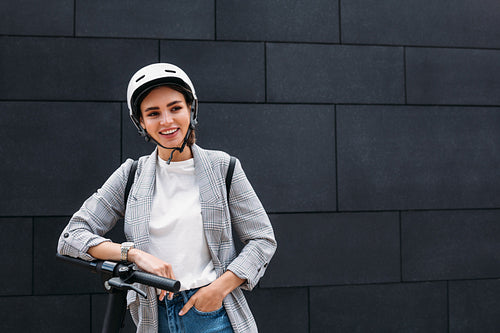 Portrait of a smiling businesswoman leaning on handlebar of electric push scooter. Young cheerful female in cycling helmet at black wall looking away .