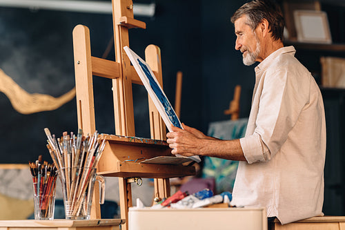 Side view of mature artist holding a canvas with picture he painted