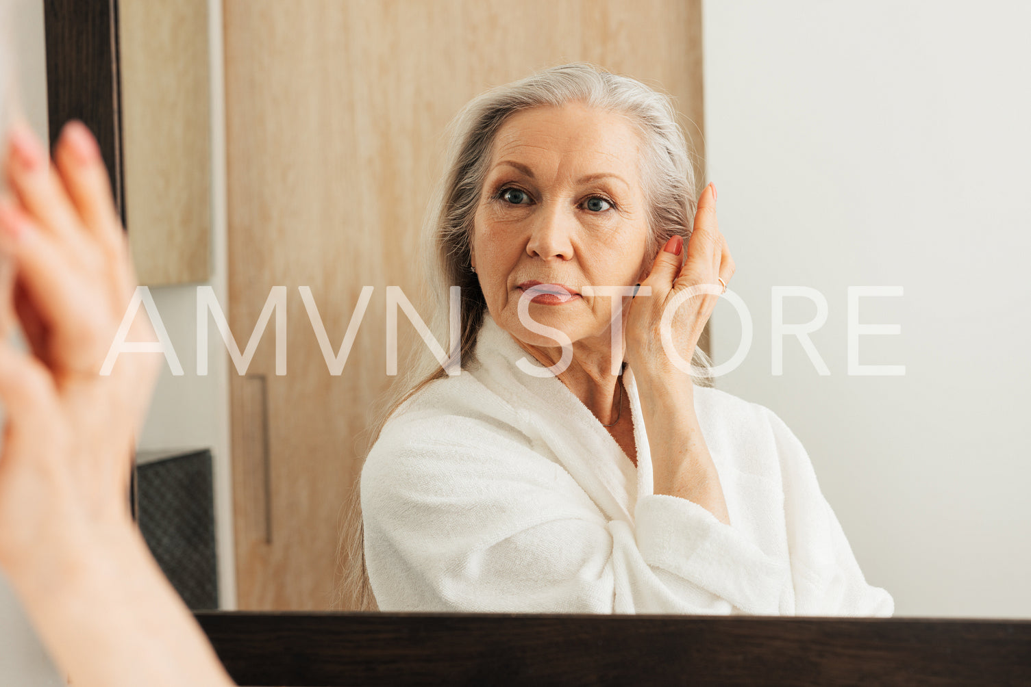 Smiling aged woman in bathrobe touching her face with fingers in bathroom