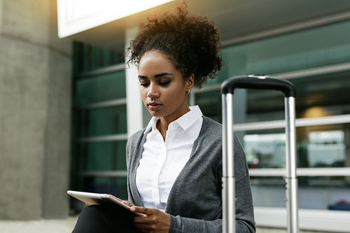 Young woman using digital tablet, sitting with suitcase at building