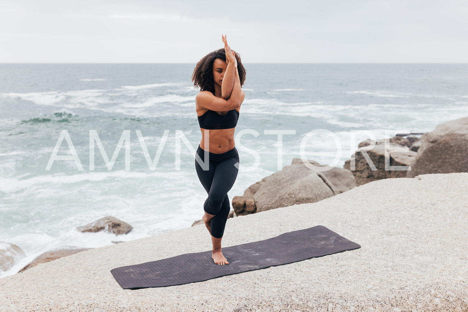 Woman standing on mat in Garudasana pose by ocean. Young female with curly hair practicing Eagle Pose.