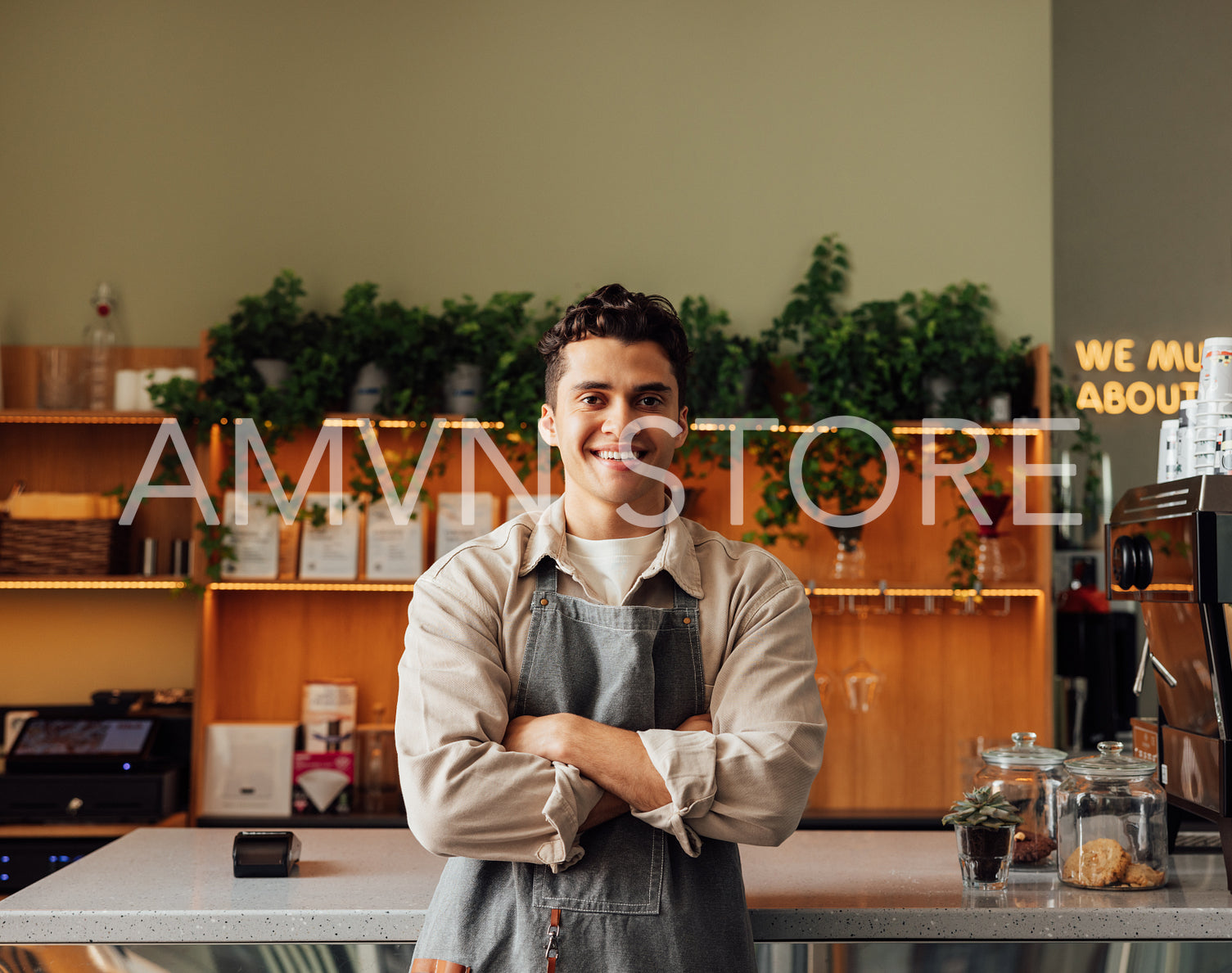 Coffee shop owner in an apron standing with crossed arms and smiling. Young handsome man leaning counter looking at a camera in a coffee shop.