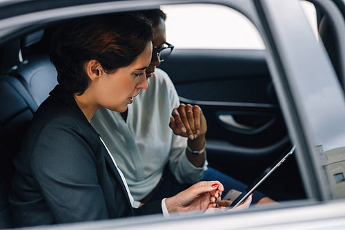 Two female colleagues looking at digital tablet and talking while sitting of a backseat of a taxi