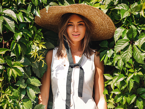 Young blond woman in big staw hat standing at wall with green leafs and looking at camera