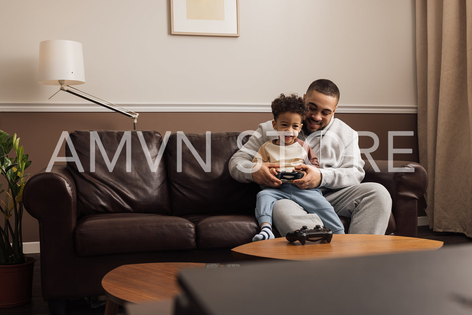 Little boy sitting on the hip of his father and holding a joystick. Male helping a kid to play a video game.