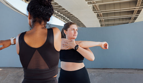 Two plus-size females in black fitness attire warming up. Young women stretch their hands under a bridge.