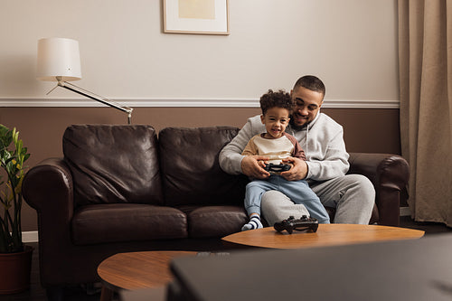 Little boy sitting on the hip of his father and holding a joystick. Male helping a kid to play a video game.