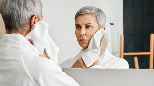 Mature woman with short grey hair looking at her reflection in bathroom mirror while wiping cheek