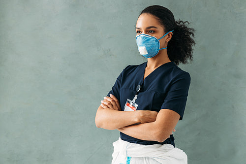 Young nurse in medical uniform with badge standing at wall with crossed arms