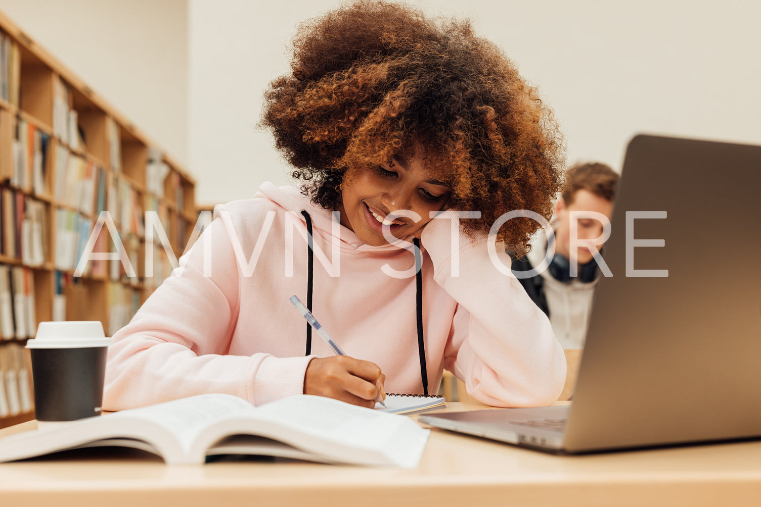 Smiling student with curly hair writing on notebook while sitting at desk in library