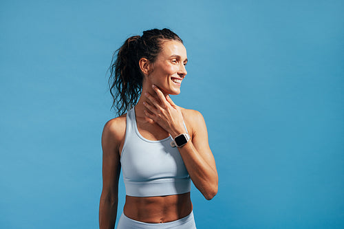 Smiling woman in fitness wear touching her neck and looking away on blue background