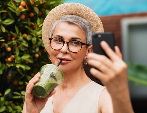 Aged woman in eyeglasses drinks smoothie