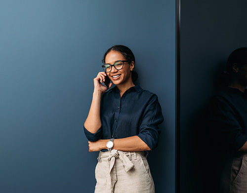 Smiling woman in formal clothes leaning blue wall talking on a mobile phone