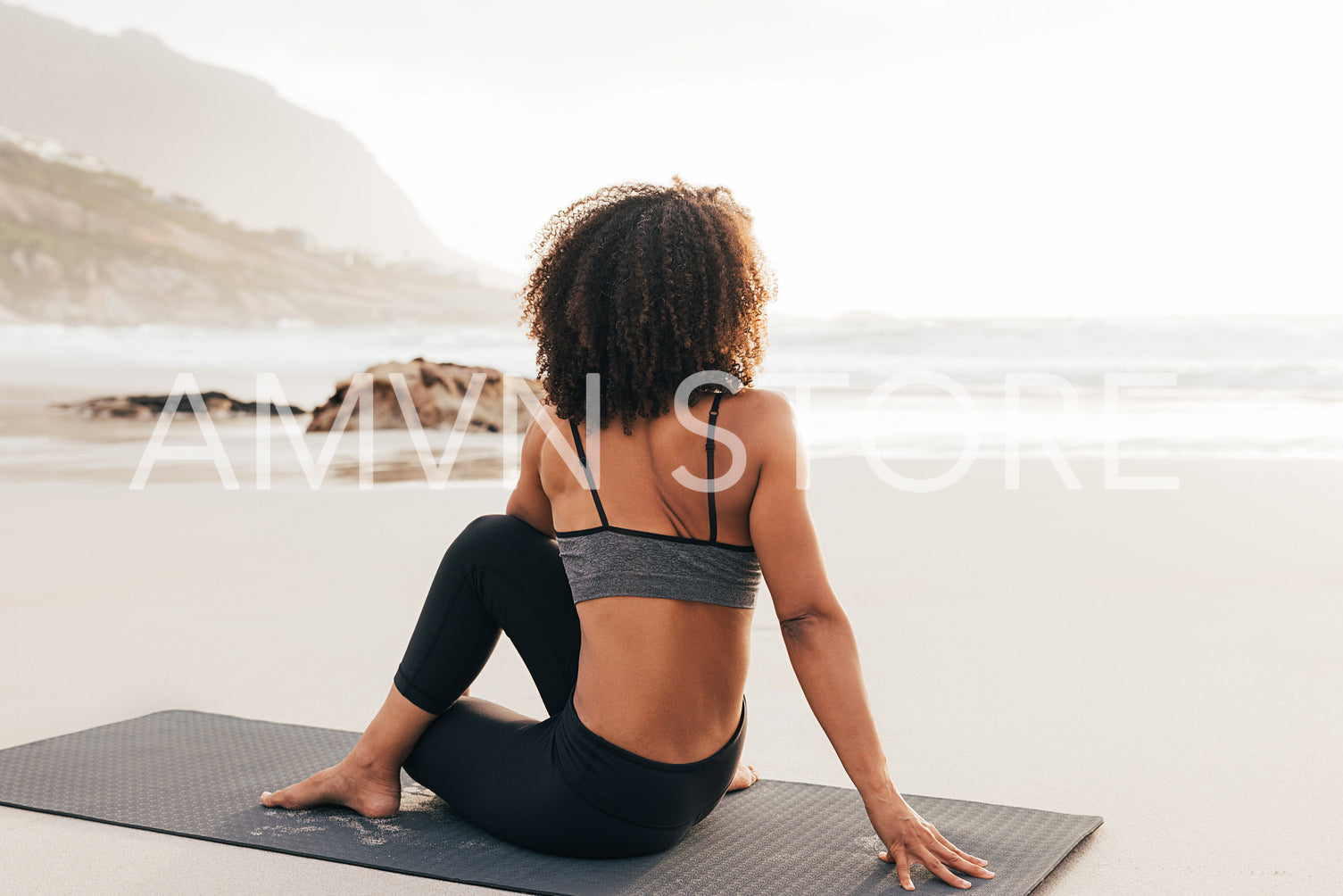 Woman sitting on a mat on beach at sunset. Back view of a female relaxing after yoga exercises.