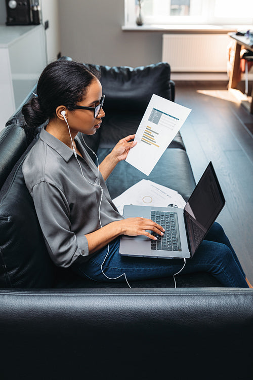 Female entrepreneur using laptop sitting on a sofa wearing earphones. Side view of a young businesswoman working from home.