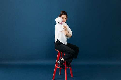 Young female in casuals sitting on a red chair in a studio with her little adorable dog