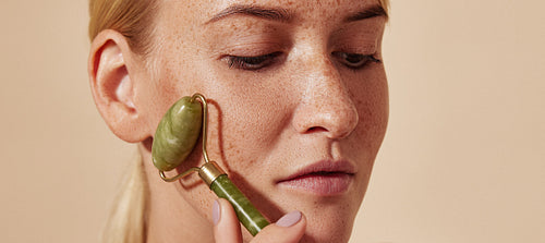 Close-up portrait of a young woman with freckles using a jade roller. Blond female doing beauty routine using face roller.