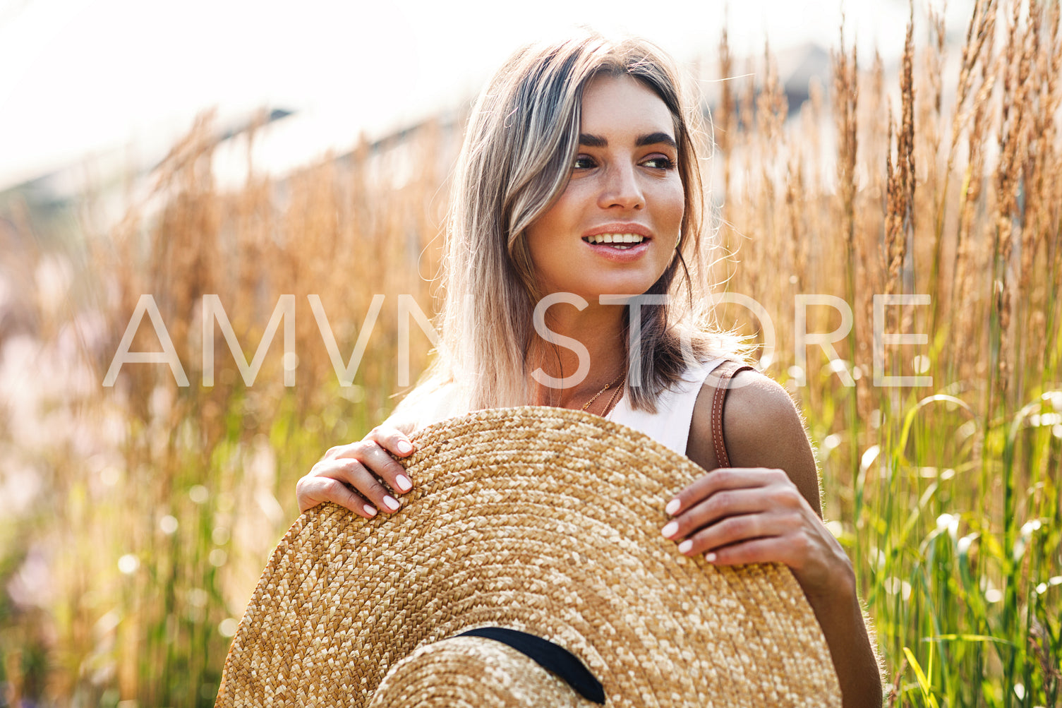 Young beautiful woman holding a big straw hat on a field surrounded by tall grass	