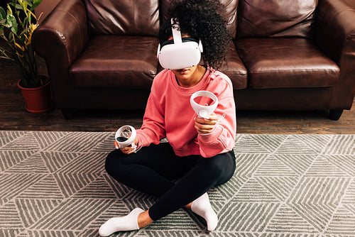 Smiling woman in living room using VR set. Young female in comfy clothes playing video games while sitting on the floor.