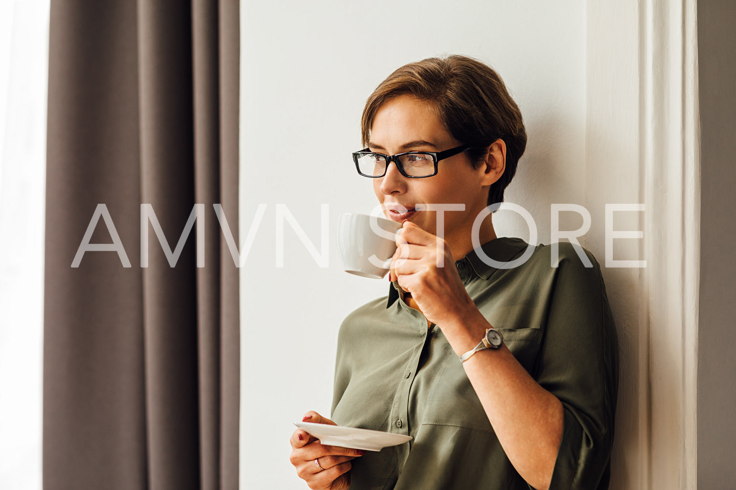 Mid adult female in formal clothes having coffee in hotel room and looking away. Business woman standing at wall in apartment and drinking coffee.	