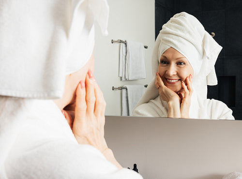 Cheerful woman admires her reflection in the bathroom mirror. Mature female touching her face with hands after the home spa.