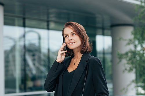 Smiling corporate person with ginger hair in black formal clothes talking on a mobile phone outdoors