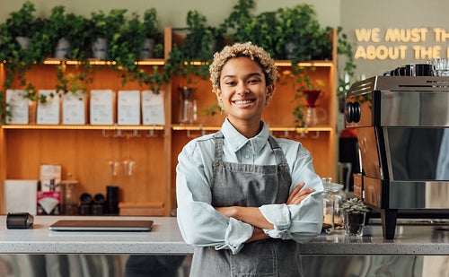 Smiling confident coffee shop owner at the counter. Female barista in apron looking at camera and smiling while leaning a counter in cafe.