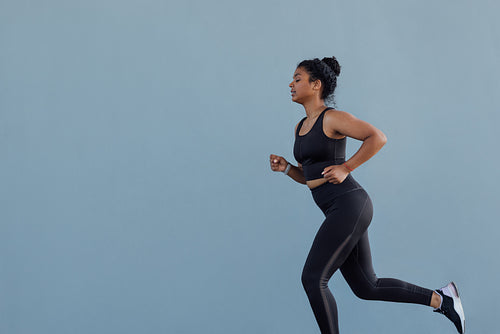 Side view of young female running at a grey wall. Woman in black fitness wear jogging outdoors.