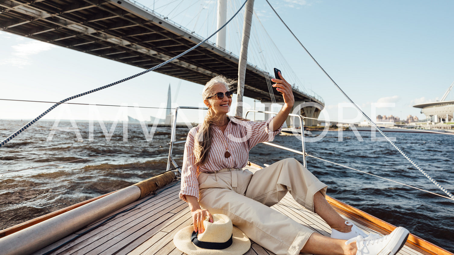 Happy mature woman on yacht trip taking a selfie. Stylish female sitting on a boat deck taking photographs on mobile phone while floating under a bridge.	