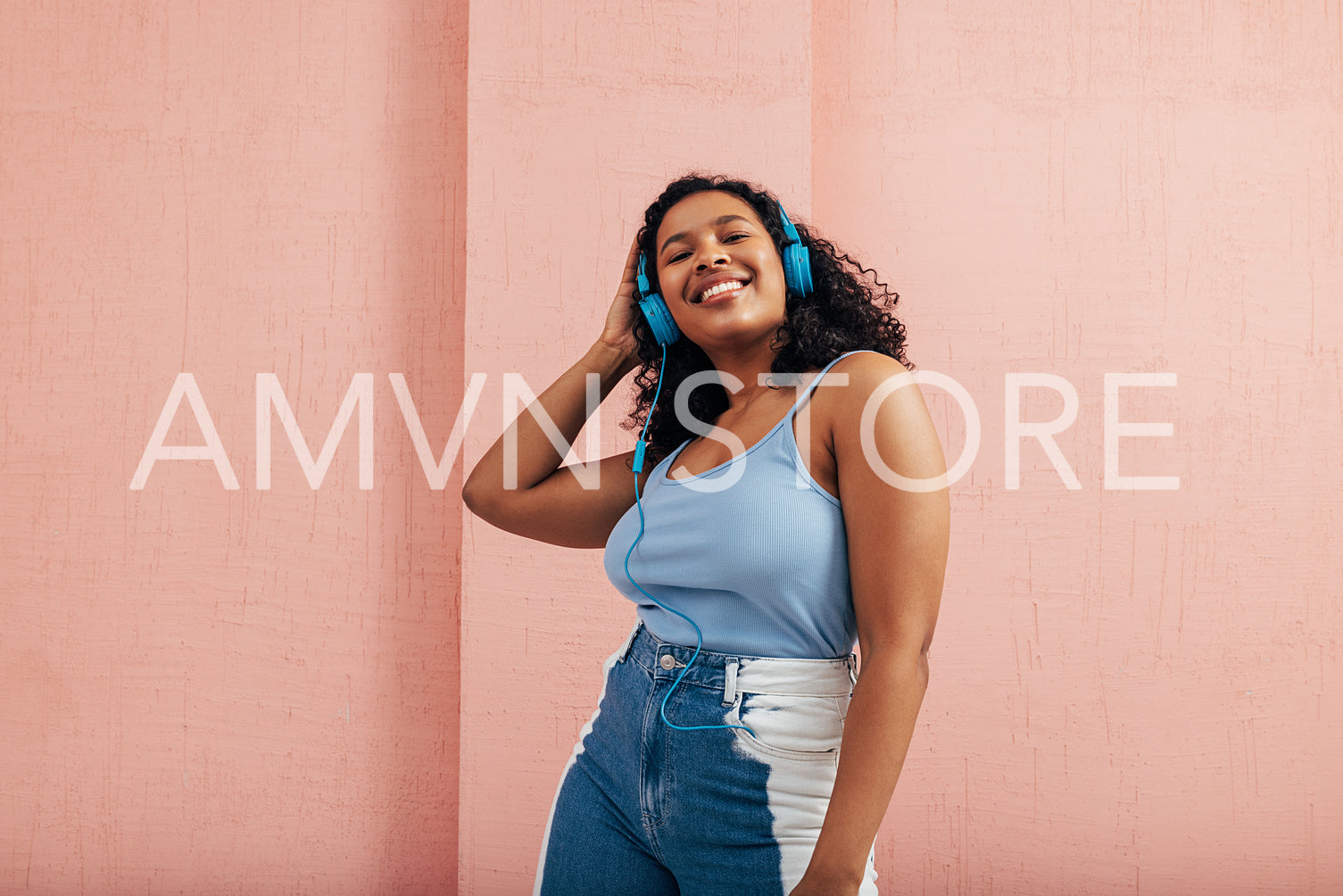 Cheerful woman with black curly hair wearing blue headphones standing at pink wall