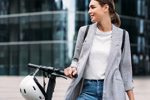 Beautiful caucasian businesswoman in formal wear holding a handlebar of electric scooter while standing against an office building