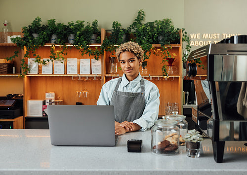 Young female entrepreneur in an apron looking at camera while standing at the counter with laptop and coffee machine