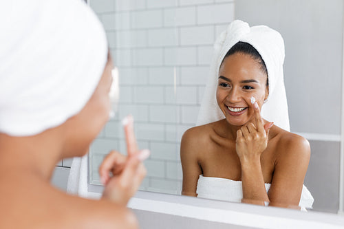 Portrait of a young smiling woman applying moisturiser to her face in the bathroom