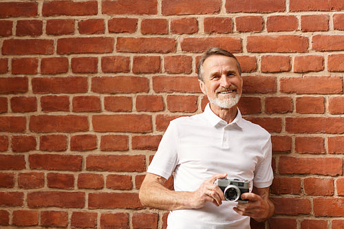 Smiling mature man looking at camera. Tourist with camera standing at brick wall.