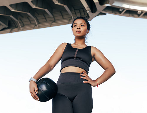 Young woman in black fitness attire under a bridge taking a break. Female athlete with medicine ball outdoors looking away.