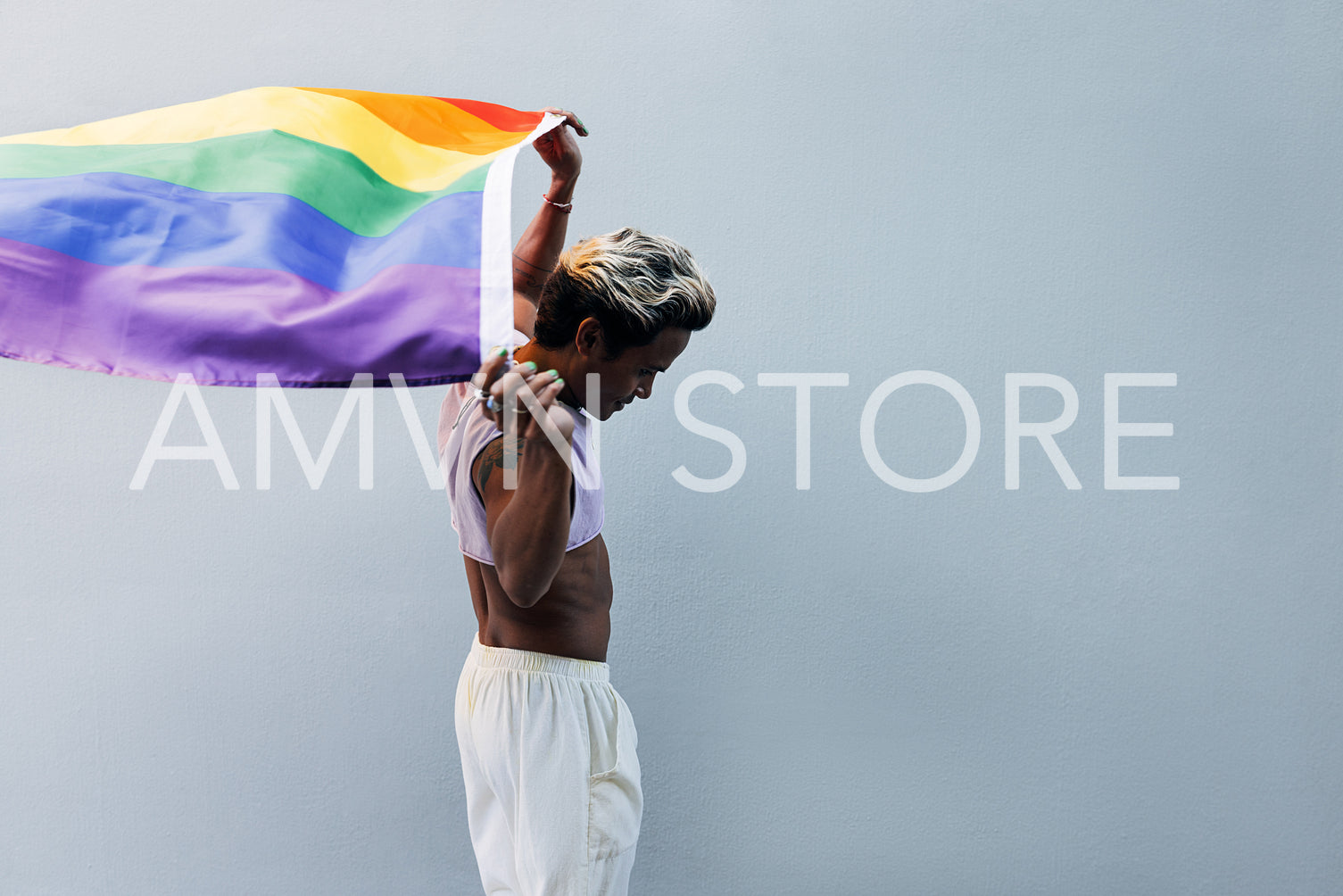 Young stylish man walking at grey wall raising hands with LGBT flag