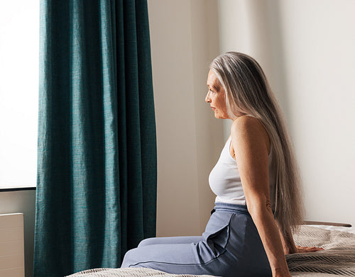 Side view of an aged woman with a long white hair sitting on a bed looking at window