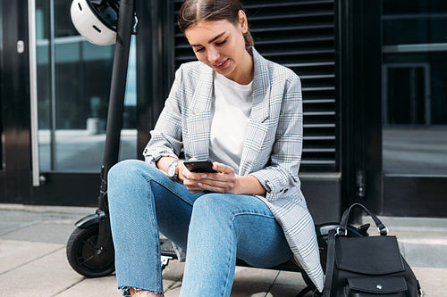 Businesswoman typing on a cell phone while sitting on electric push scooter at building