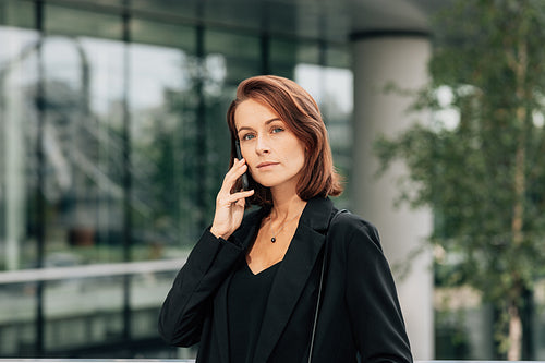 Confident businesswoman with ginger hair talking on a mobile phone. Middle aged female in black formal clothes talking on a cell phone outdoors.