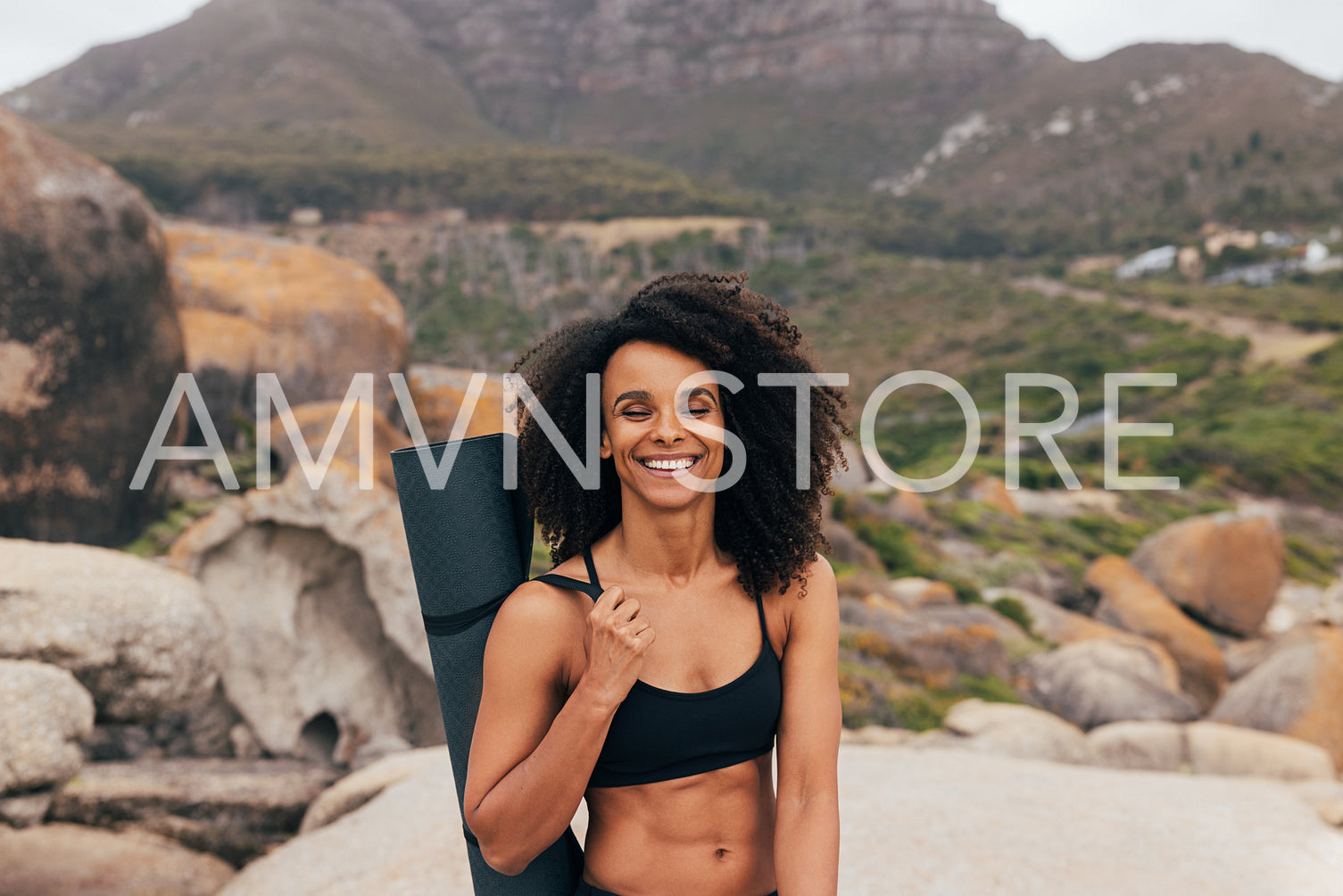 Portrait of a smiling fit woman with closed eyes holding a mat while standing by mountains in valley