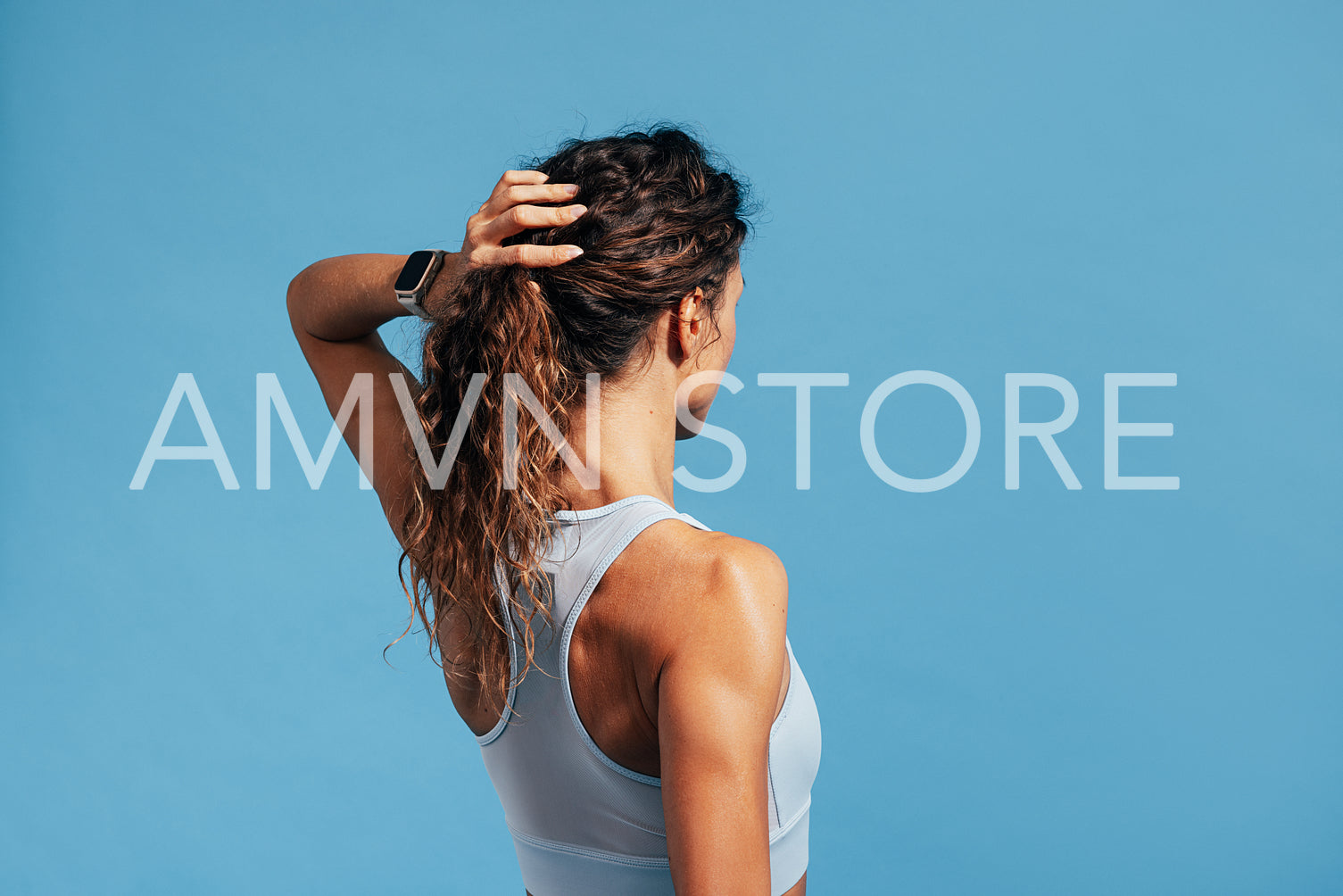 Back view of a muscular woman adjusting her hair while standing on blue background